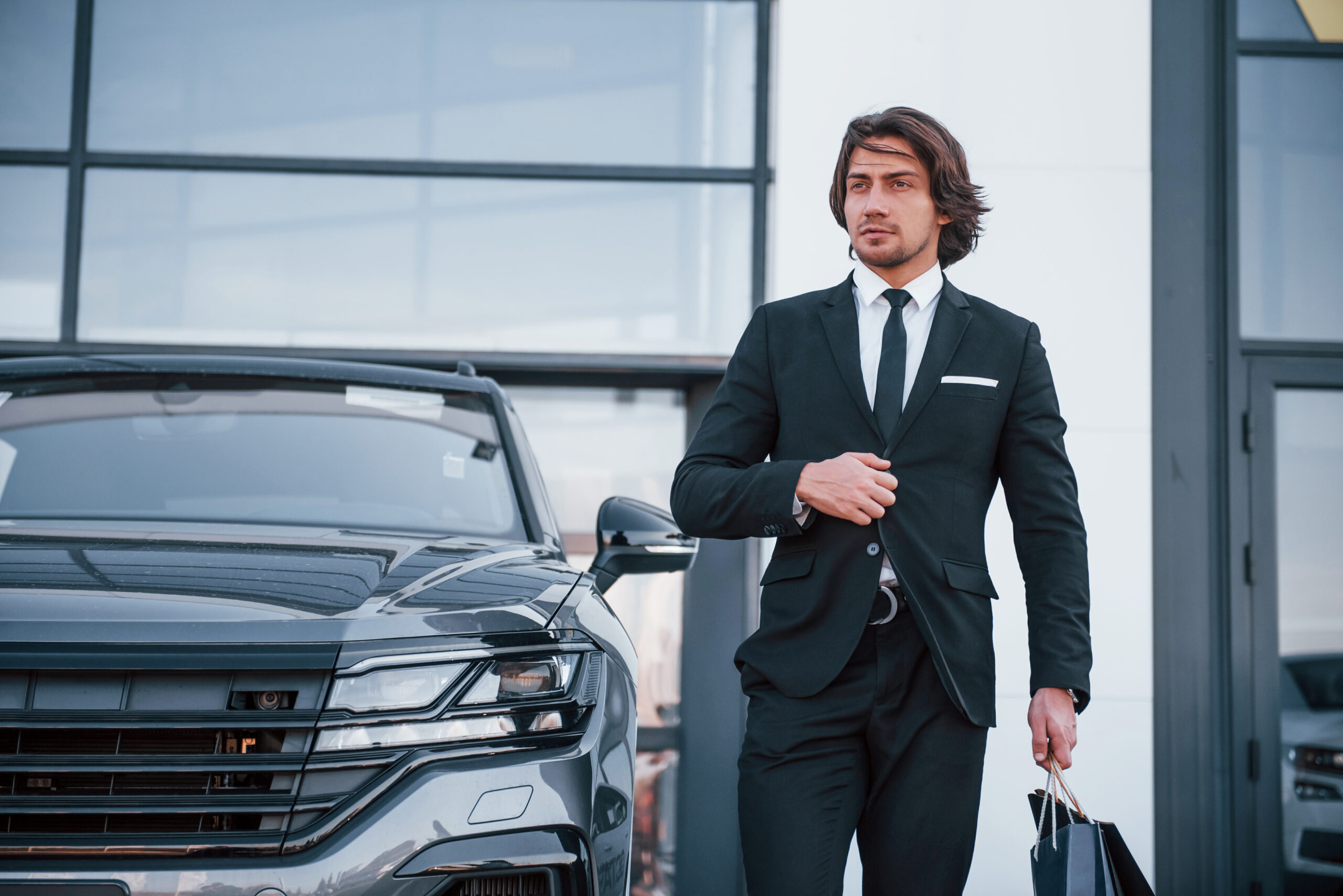 Portrait of handsome young businessman in black suit and tie outdoors near modern car and with shopping bags